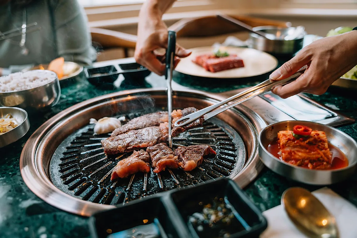 Sliced beef grilling on a Korean barbecue grill beside kimchi, rice and side dishes in stainless steel bowls