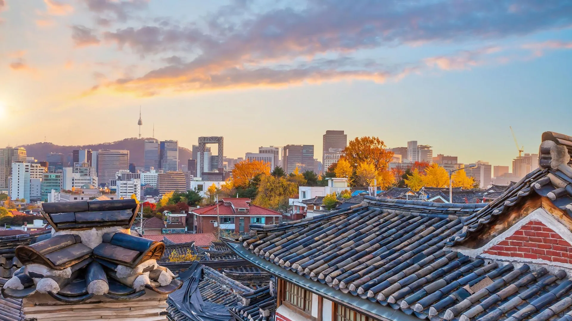 Bukchon tiled roofs and autumn trees in front of the Seoul skyline, with N Seoul Tower on the ridge beneath golden evening light