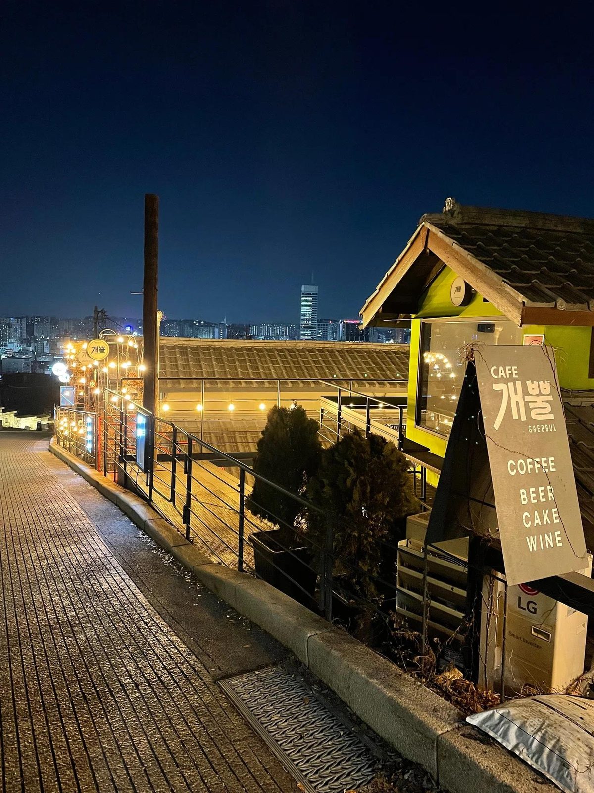 Café sign and string lights on a hillside street in Ihwa-dong at night with the Seoul skyline in the distance