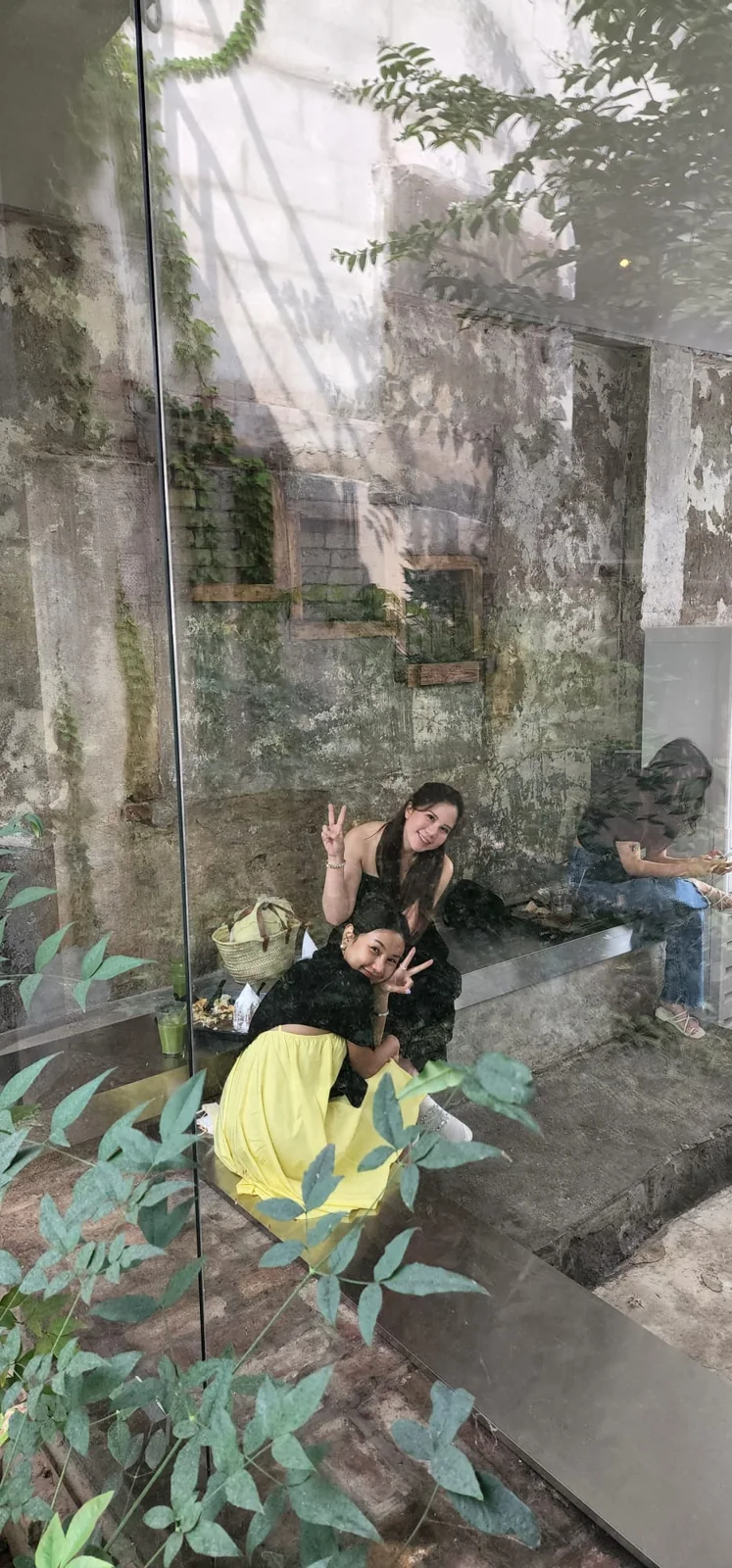 Two smiling travelers making peace signs at a Seongsu-dong cafe, seen through a glass wall with leafy plants and weathered concrete walls