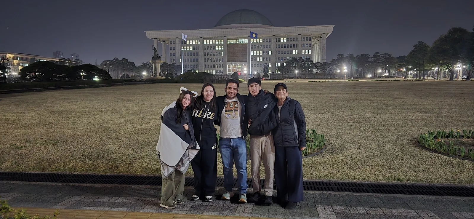 Travelers posing after dark on the grounds of Korea's National Assembly, its colonnaded facade, green dome and flags lit behind them
