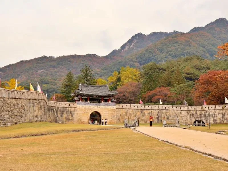 Stone gateway and wall with a grassy foreground and autumn-coloured mountain ridges at Mungyeong Saejae