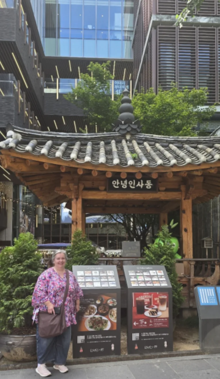 A traveler smiling in front of a traditional wooden pavilion with a grey tiled roof on a Insadong street, with a modern building and shop directory boards behind