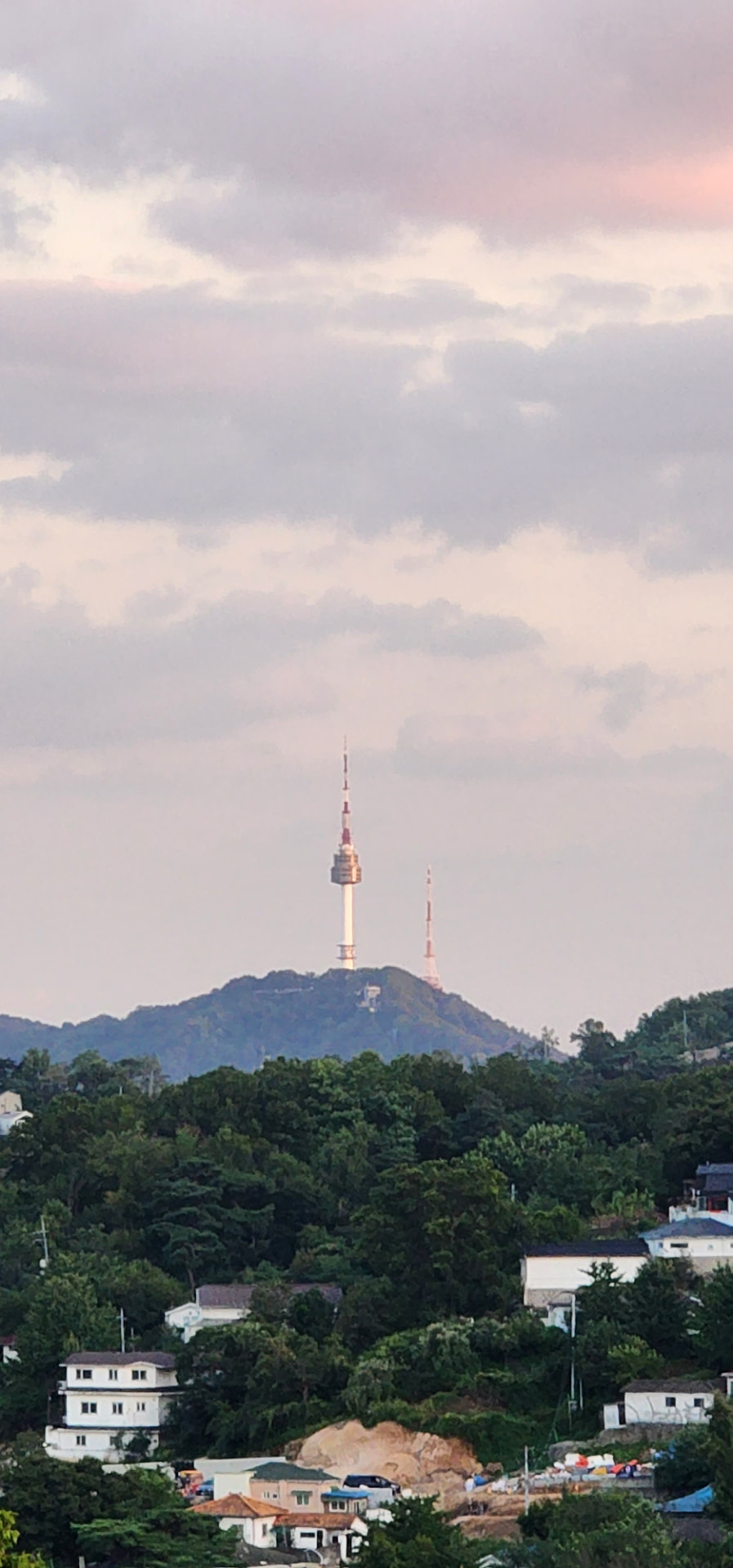 N Seoul Tower rising above forested hills and hillside homes beneath soft sunset clouds