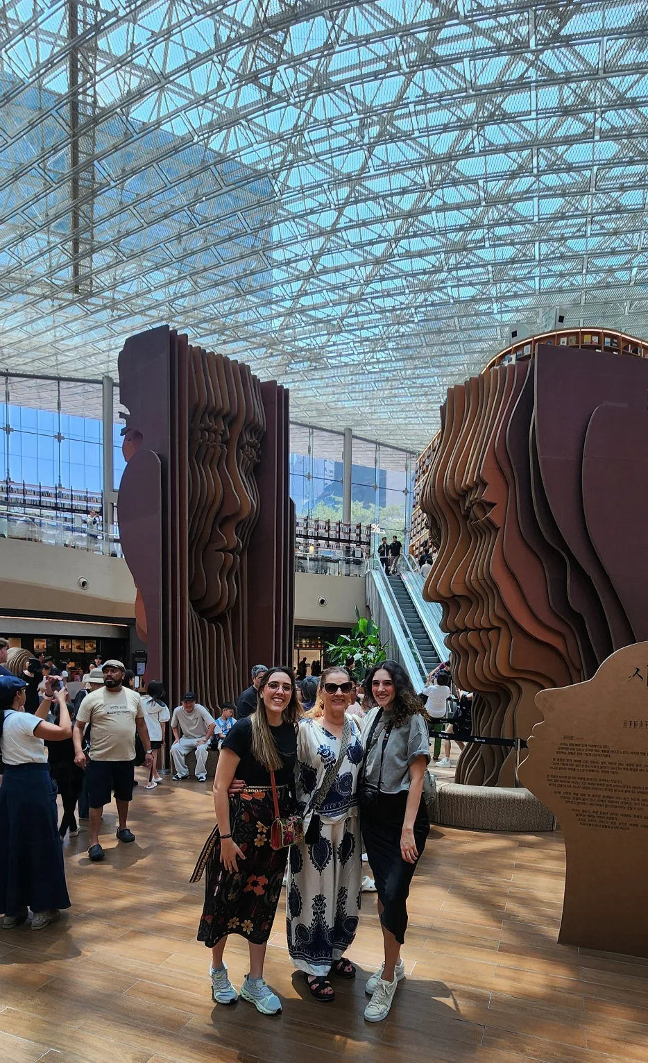 Three travelers smiling in the glass-roofed atrium of the Starfield Library at COEX, between tall bookshelves and two large head-shaped sculptures