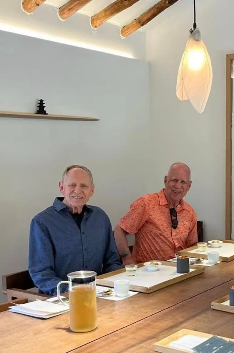 Two smiling travelers seated at a long wooden table in a modern Hanok-style Korean restaurant, with wooden trays, small dishes and a glass pitcher of iced tea in front of them