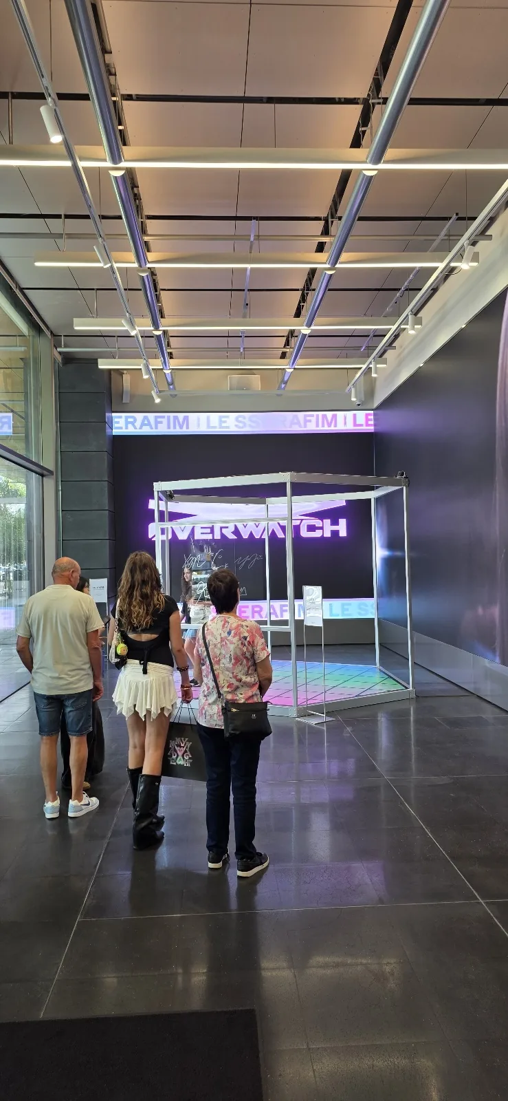 Travelers standing in a modern K-pop agency lobby beside a large media screen and a glass photo booth