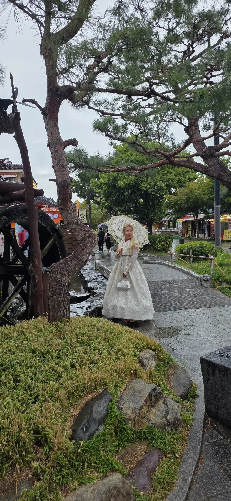 A traveler in a white modern hanbok holding a lace parasol on a stone bridge beside a stream with a wooden water wheel, with traditional Hanok rooftops behind