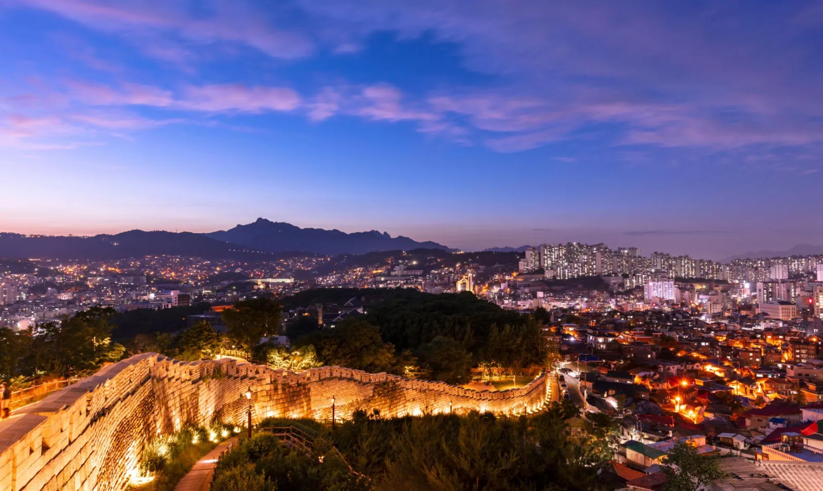 Illuminated Seoul City Wall curving along a ridge at dusk above the glittering city lights, with pink evening clouds and a mountain silhouette behind