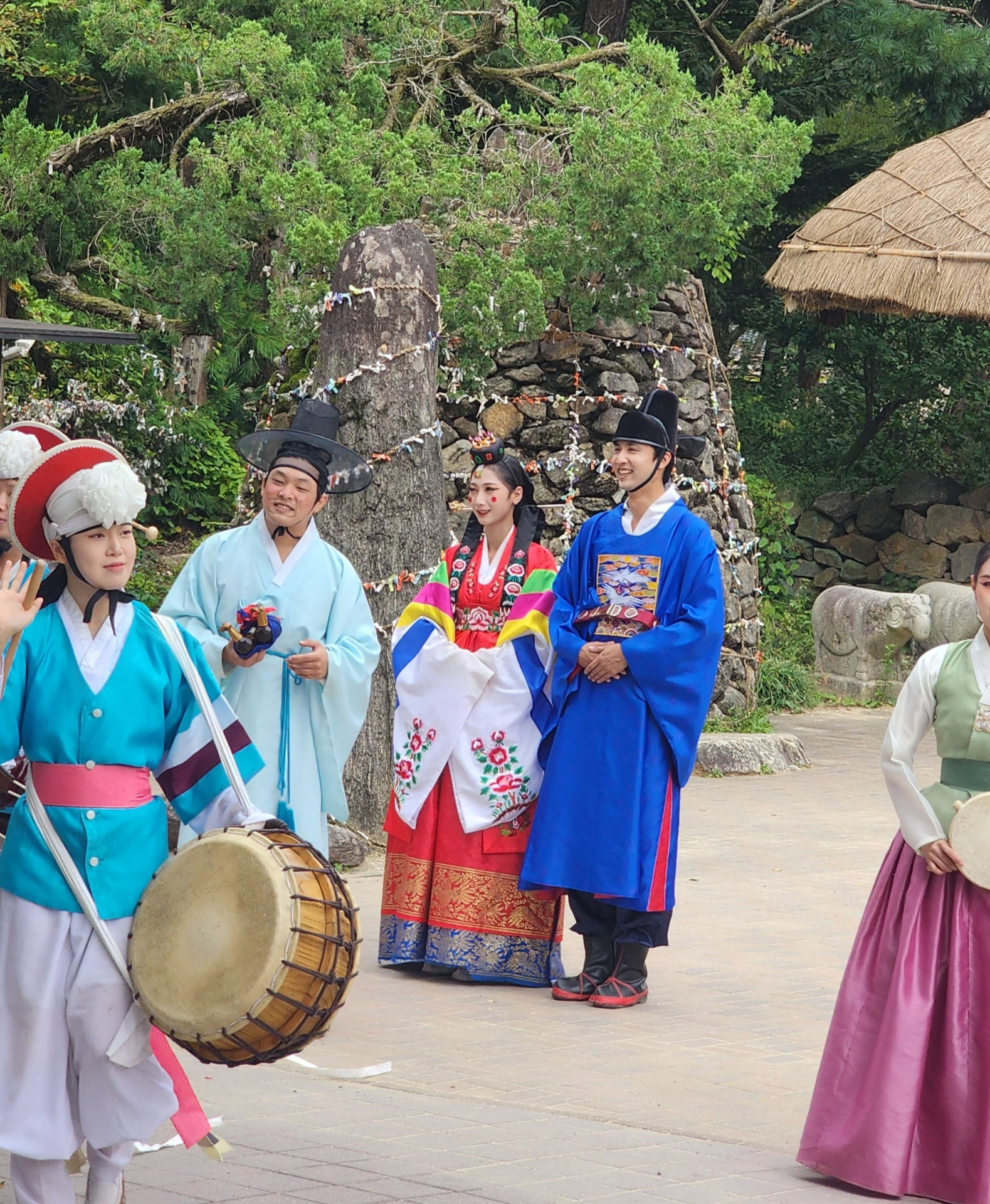 Nongak performers in hanbok with drums and ribbon hats beside a stone tower and thatched pavilion