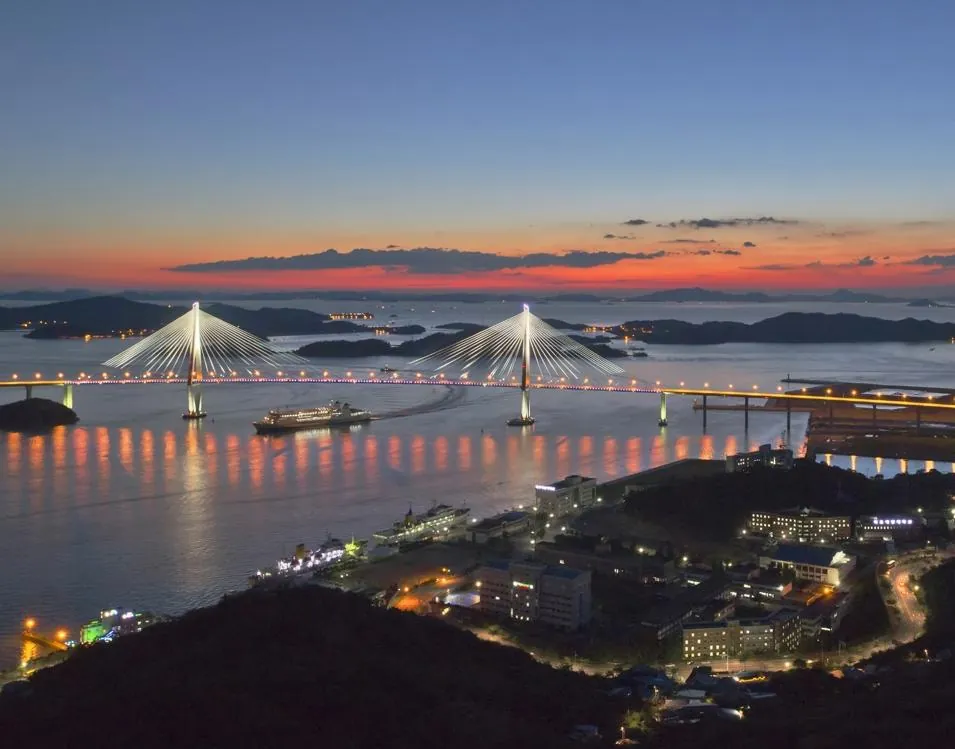 Mokpo Bridge lit at dusk over the harbour with a ferry passing beneath and sunset along the coast