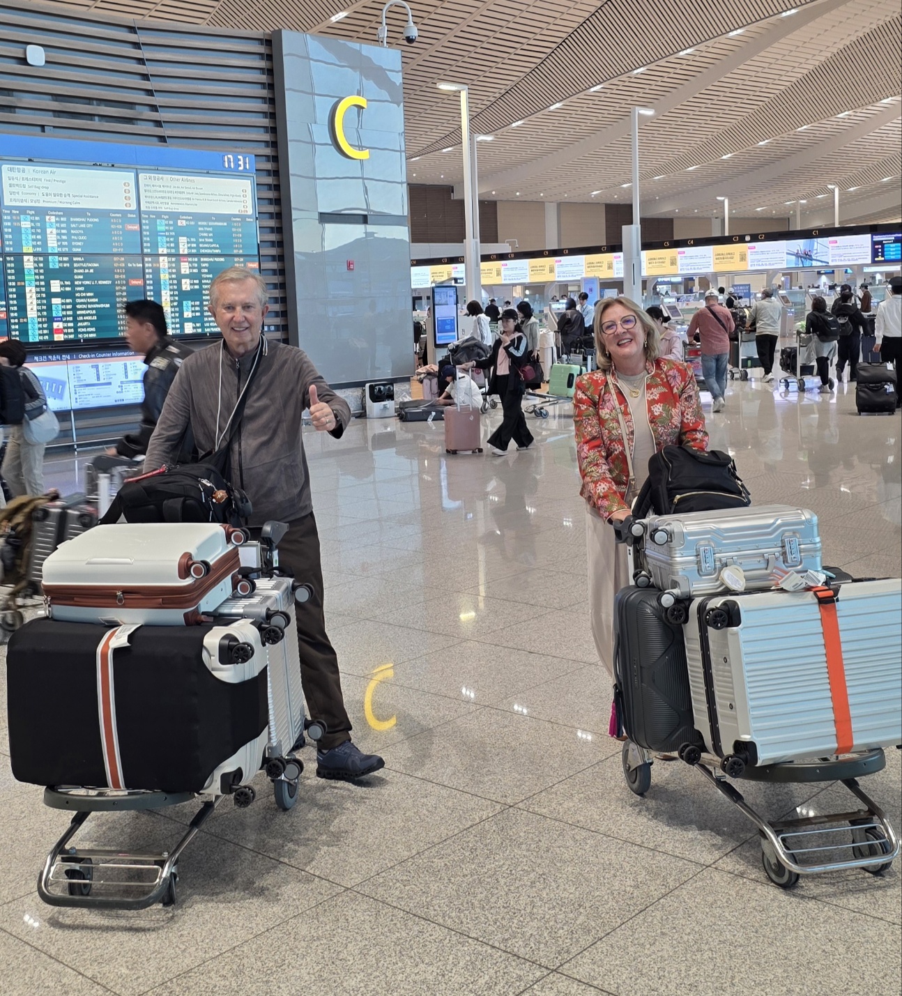 A Brazilian couple smiling with luggage carts in the arrivals hall of Incheon International Airport, a flight information board behind them