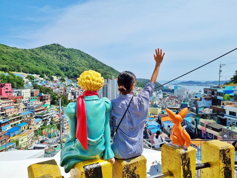 Visitor waving beside the Little Prince statue overlooking colourful Gamcheon Culture Village