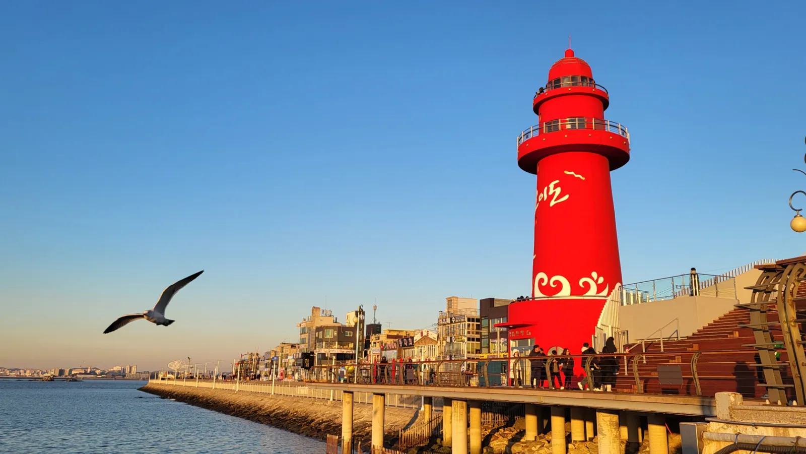 Bright red Oido Lighthouse beside the waterfront promenade under a clear blue sky