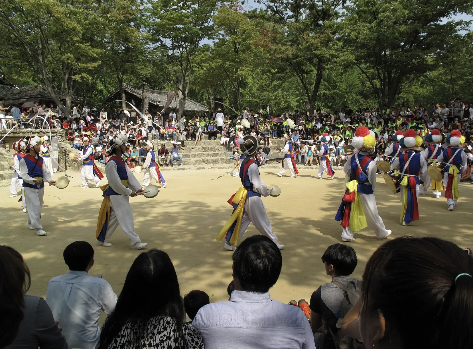 Nongak performers in white costumes with colourful sashes and ribbon hats playing hand drums in the outdoor arena at Korean Folk Village
