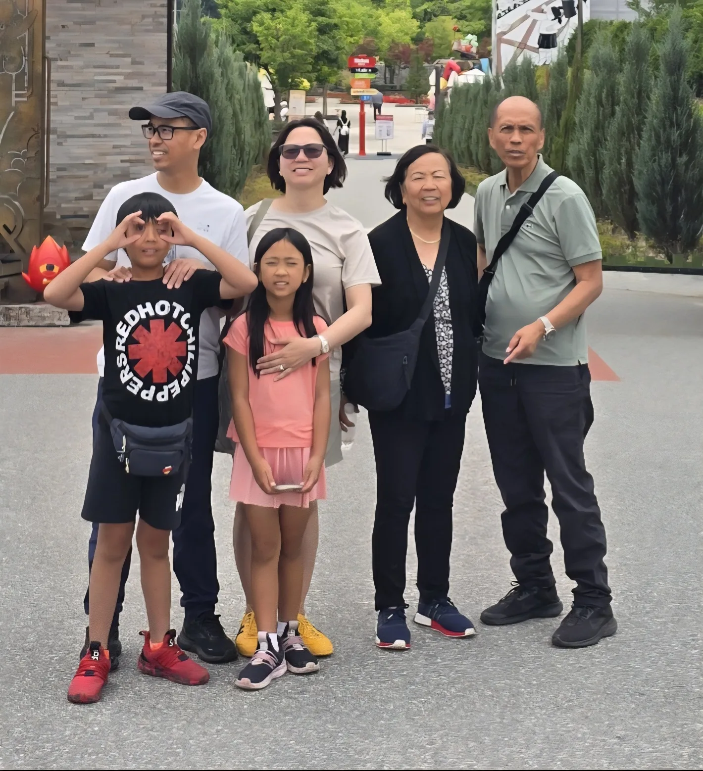 Six travelers of different generations posing together on a tree-lined paved walkway, with a red character sculpture and colorful signposts behind them