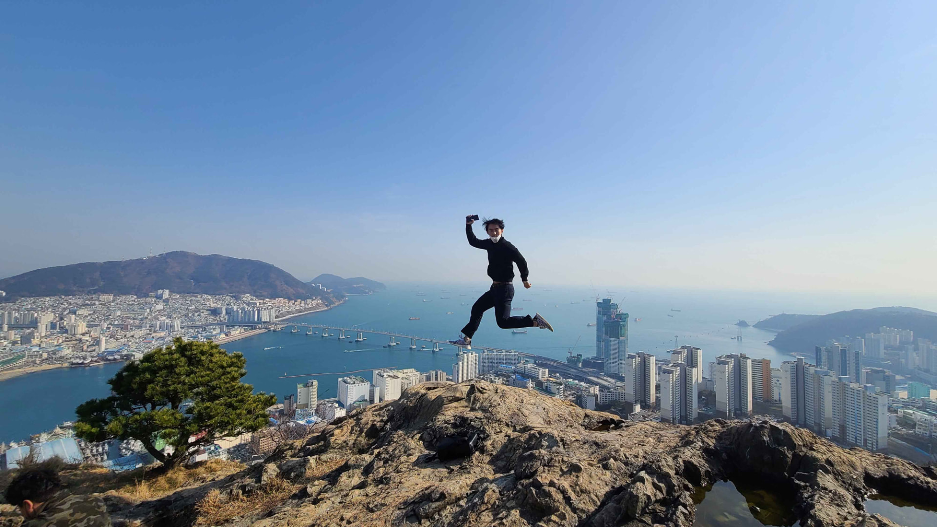 Traveller leaping joyfully on a rocky hilltop overlooking Busan's harbour and city skyline