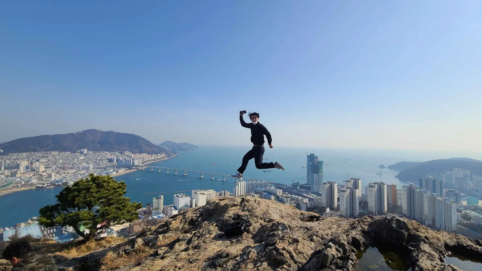 Traveller leaping joyfully on a rocky hilltop overlooking Busan's harbour and city skyline