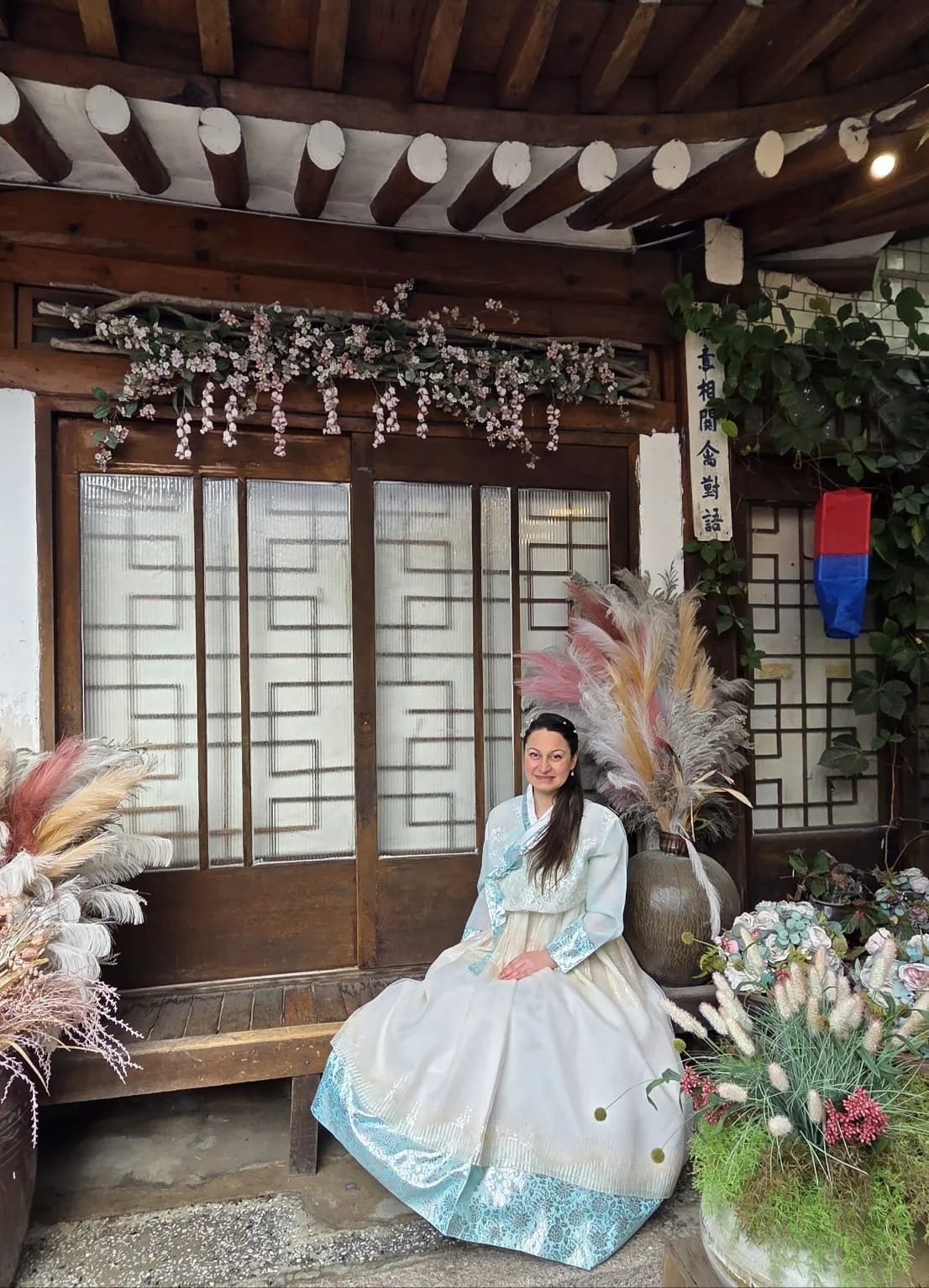 A traveler in a white and light-blue hanbok sitting on the wooden bench of a traditional hanok tea house decorated with dried flowers and a Korean sign