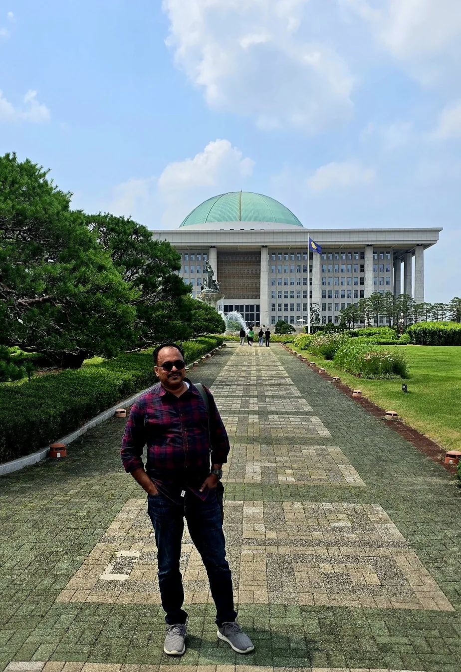 A traveler standing on a tree-lined path with the green dome of the National Assembly building behind