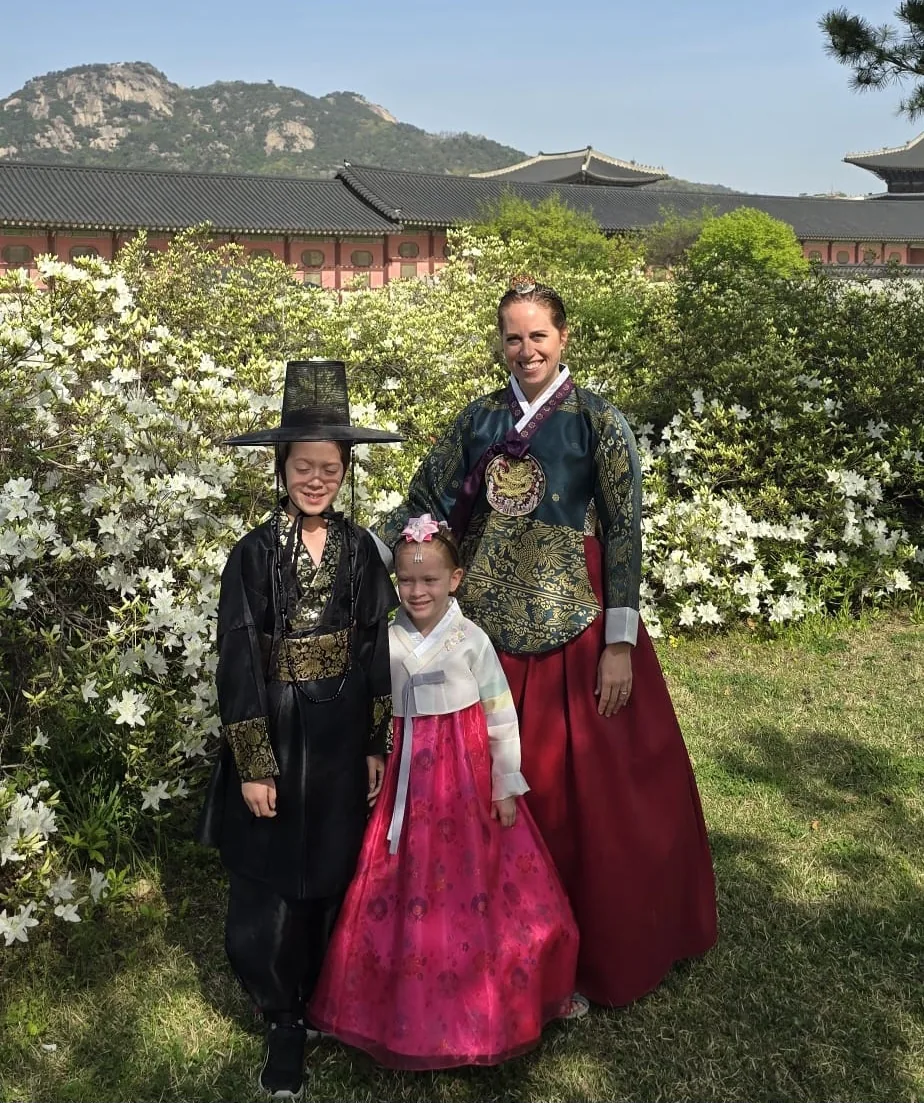 Three travelers wearing colorful hanbok beside flowering shrubs and a palace pavilion