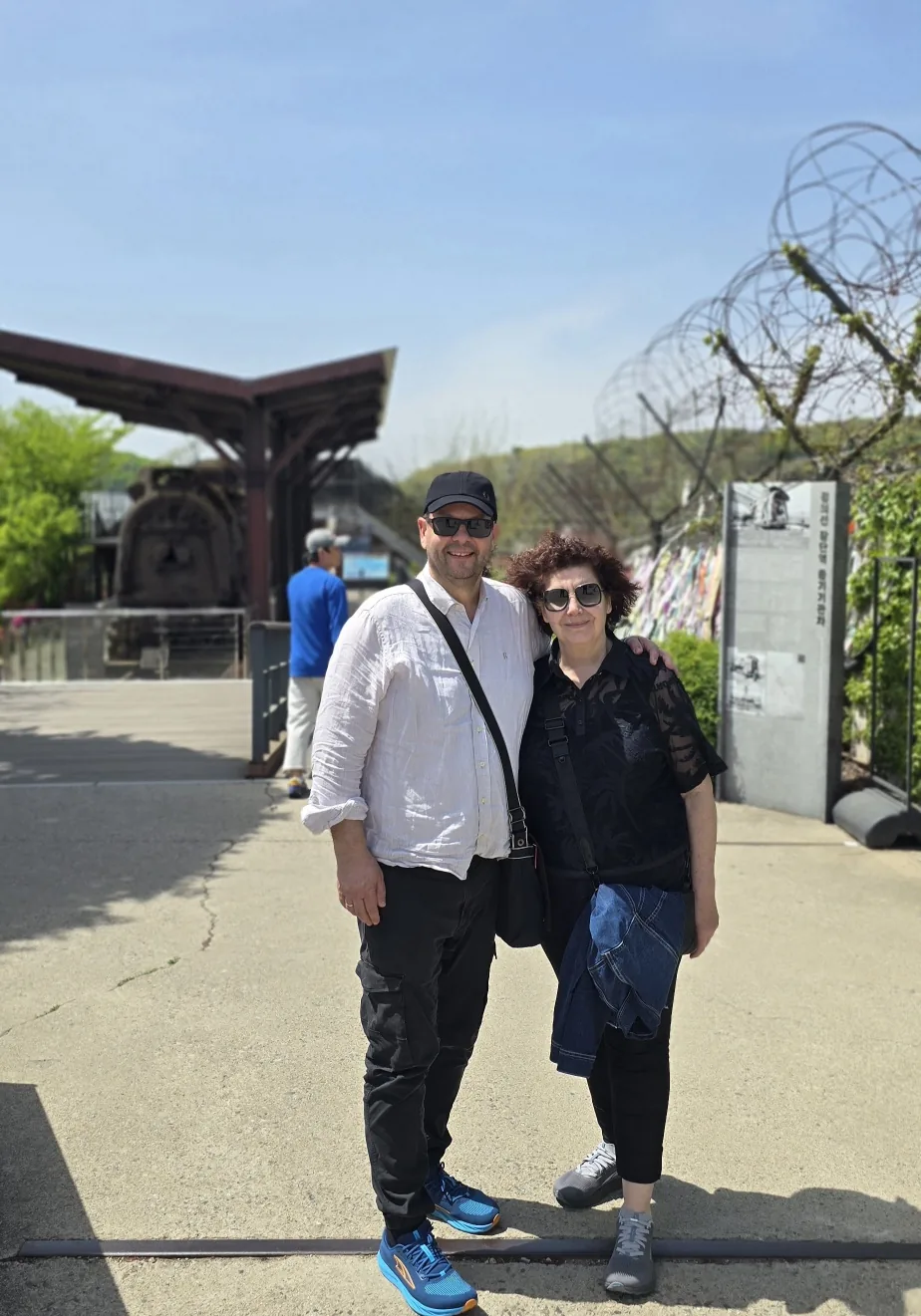 Two travelers posing beside the preserved Jangdan Station steam locomotive and its information sign at Imjingak, with a barbed-wire security fence behind